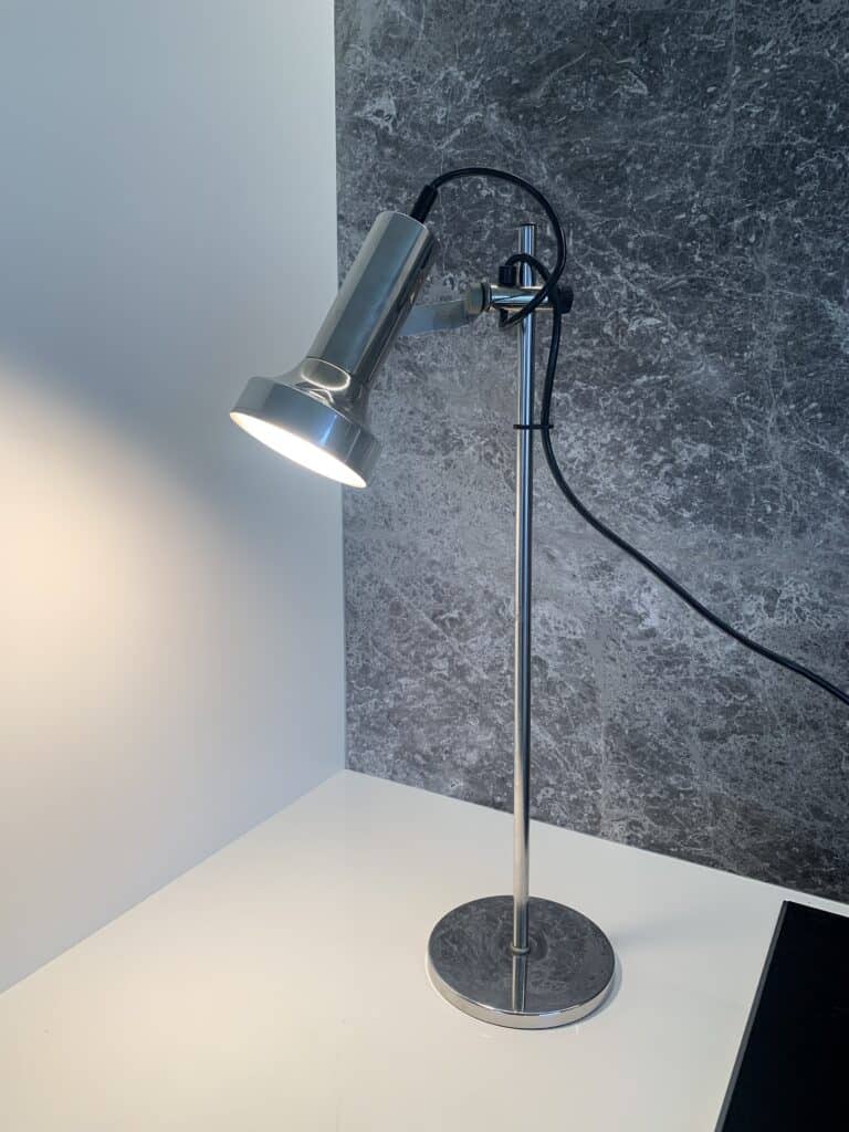 Een moderne, metalen bureaulamp met een ronde voet staat op een wit oppervlak en werpt licht op de muur. De achtergrond heeft een getextureerd, donkergrijs oppervlak en er is een zwart snoer zichtbaar.