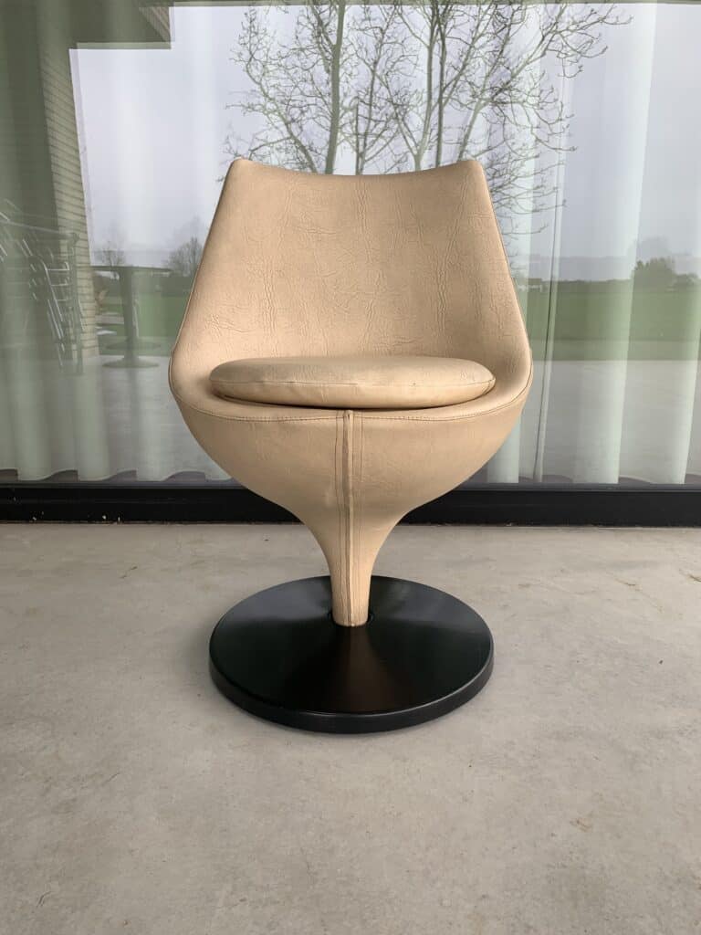 Een beige, moderne draaistoel met een gebogen rugleuning en een zitting op een zwarte voet, geplaatst op een betonnen vloer voor grote glazen ramen met transparante gordijnen en uitzicht naar buiten.