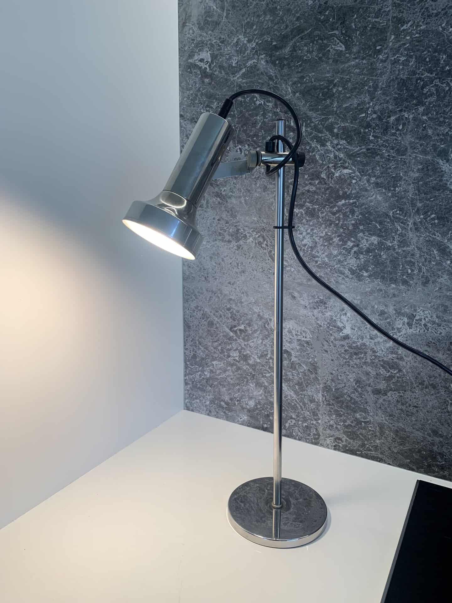 Een moderne, metalen bureaulamp met een ronde voet staat op een wit oppervlak en werpt licht op de muur. De achtergrond heeft een getextureerd, donkergrijs oppervlak en er is een zwart snoer zichtbaar.