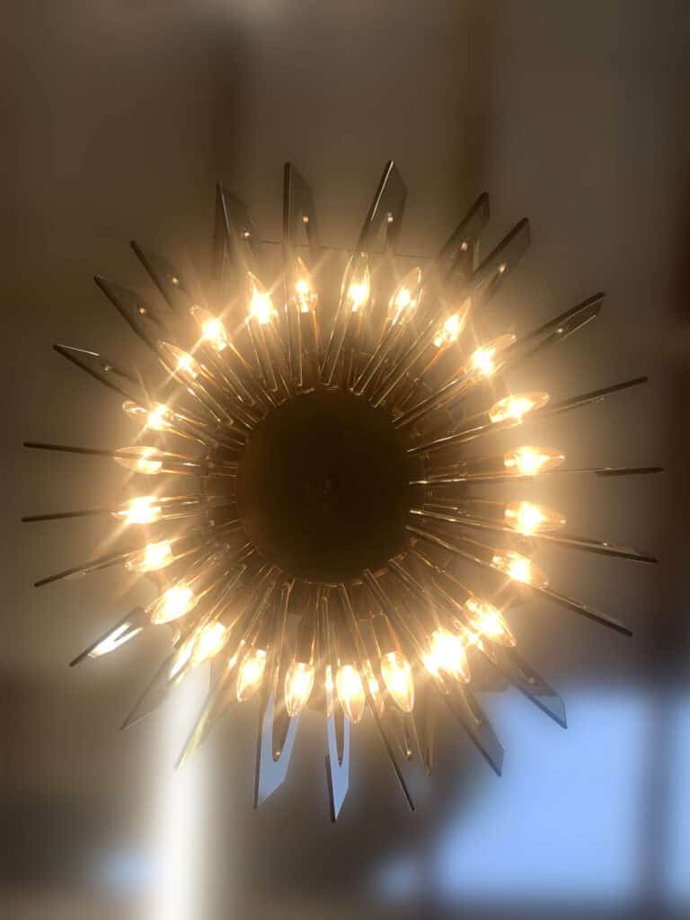 Een ronde kroonluchter met talloze brandende gloeilampen en metalen spaken die naar buiten stralen, waardoor het lijkt op een zonnestraal. Gefotografeerd van onderaf tegen een onscherpe achtergrond van het plafond.