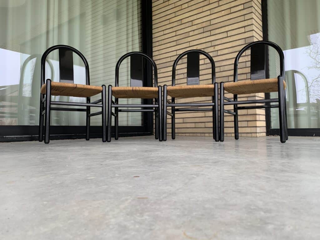 Vier stoelen met zwarte frames en gevlochten zittingen staan op een rij op een betonnen vloer buiten een gebouw met grote glazen ramen en beige bakstenen muren.