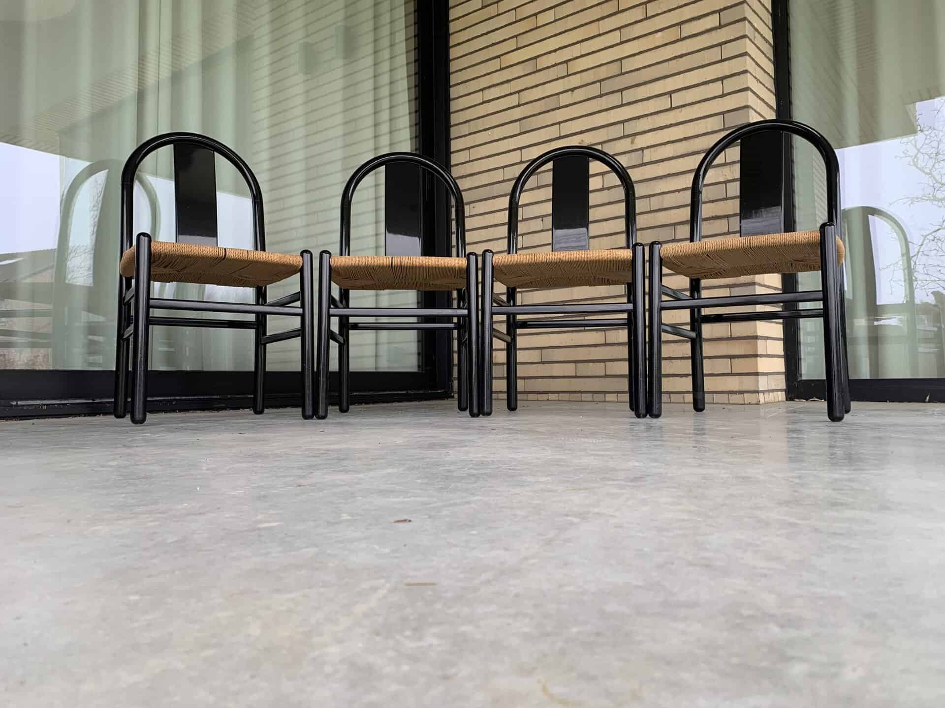 Vier stoelen met zwarte frames en gevlochten zittingen staan op een rij op een betonnen vloer buiten een gebouw met grote glazen ramen en beige bakstenen muren.