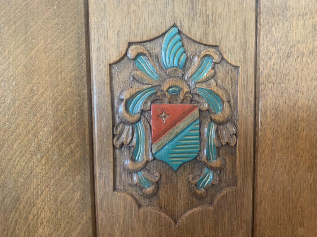Een houten paneel met een gesneden en geschilderd wapenschild met een rood en blauwgroen schild, gouden omtreklijnen en decoratieve bladachtige ontwerpen in blauwgroen en goud boven en rond het schild.