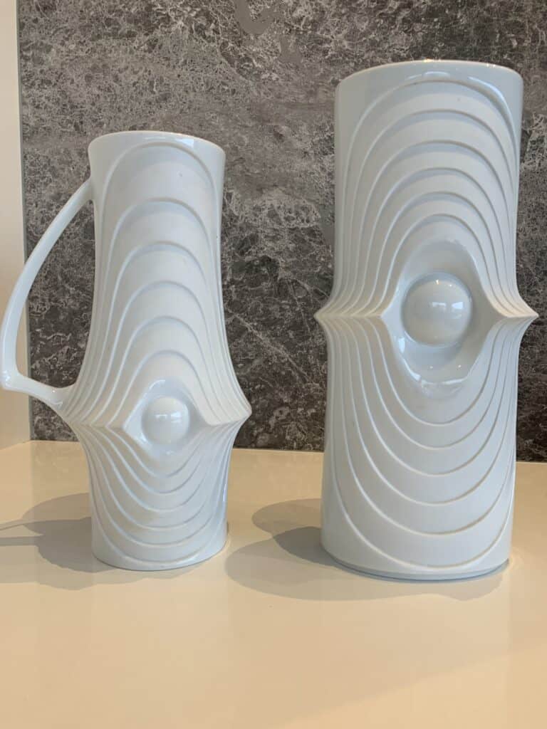 Twee hoge, witte keramische potten met verhoogde, concentrische ovale patronen en een centrale uitstulping op elk; de ene is een kan met een handvat, de andere een cilindrische vaas. Ze staan op een wit oppervlak tegen een grijze, gemarmerde achtergrond.
