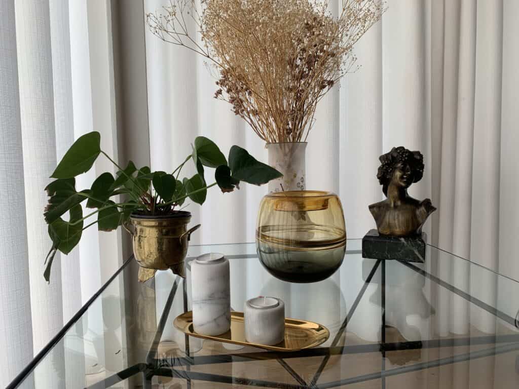 Een glazen tafel met een potplant, marmeren kaarsenhouders op een gouden dienblad, een glazen vaas met gouden accenten, een vaas met gedroogde bloemen en een klein bustebeeldje, dit alles voor witte gordijnen.