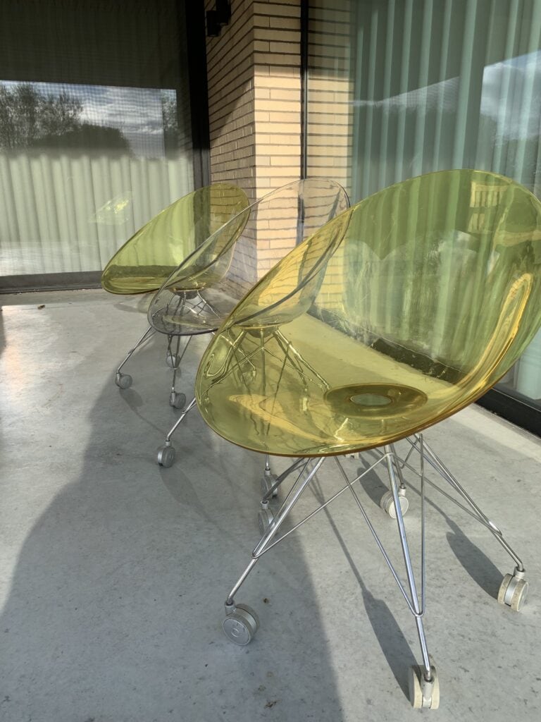 Drie moderne, doorschijnende gele stoelen met metalen frames en wielen staan op een rij op een betonnen terras buiten een gebouw met grote ramen en lichte bakstenen muren.