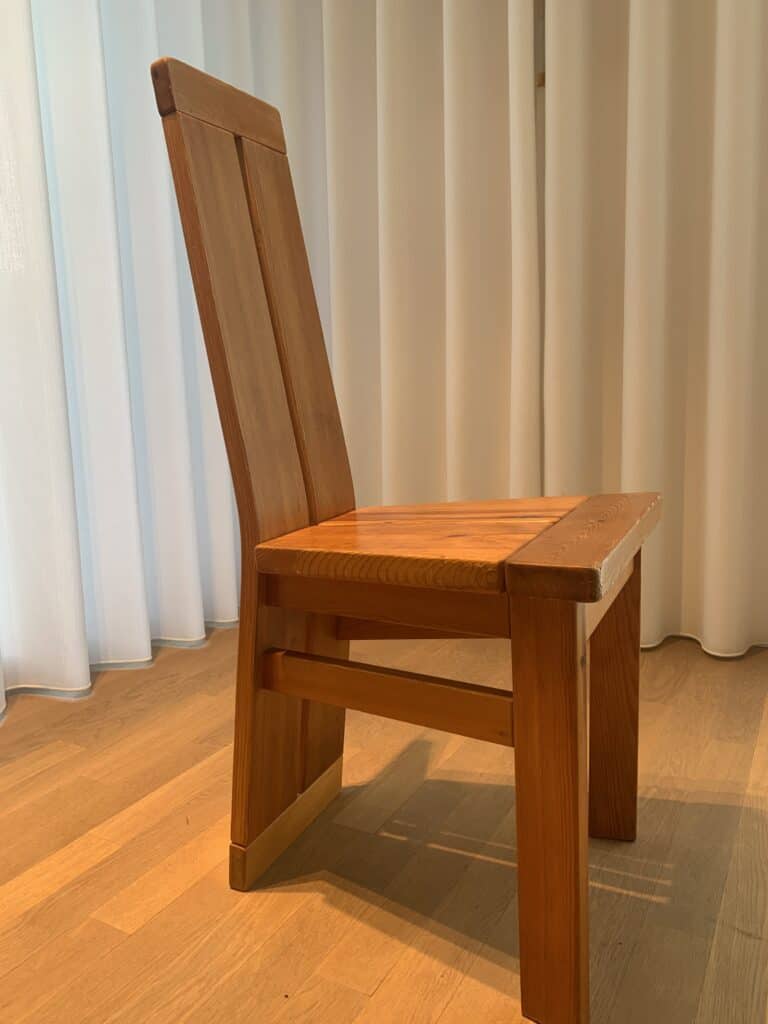 Een houten stoel met een hoge, schuine rugleuning en stevige poten staat op een lichte houten vloer, voor witte gordijnen die tot aan de vloer reiken.