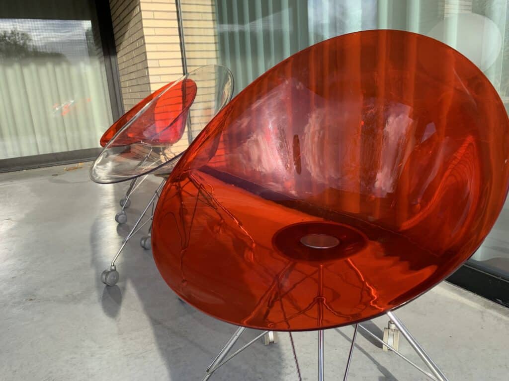 Twee moderne, ronde stoelen met doorschijnende plastic zittingen – één rood en één doorzichtig – staan op een betonnen terras, vlakbij grote glazen deuren met verticale jaloezieën en een bakstenen muur op de achtergrond.