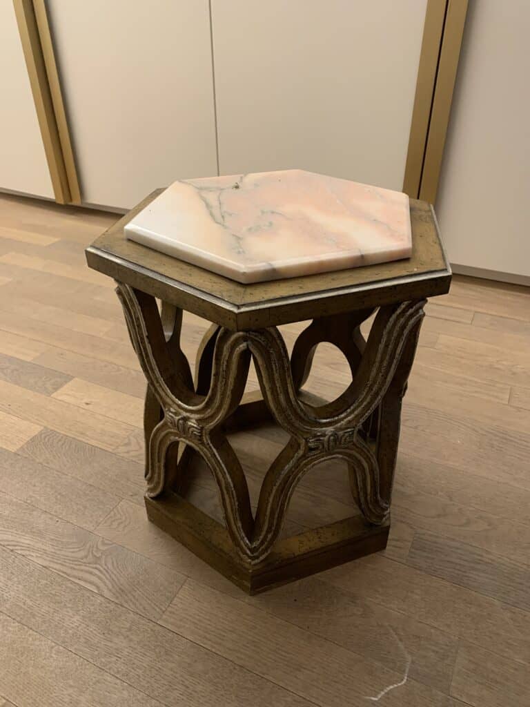 Een klein zeshoekig bijzettafeltje met een sierlijke, gesneden houten basis en een zeshoekig roze en wit marmeren blad, geplaatst op een houten vloer binnenshuis.