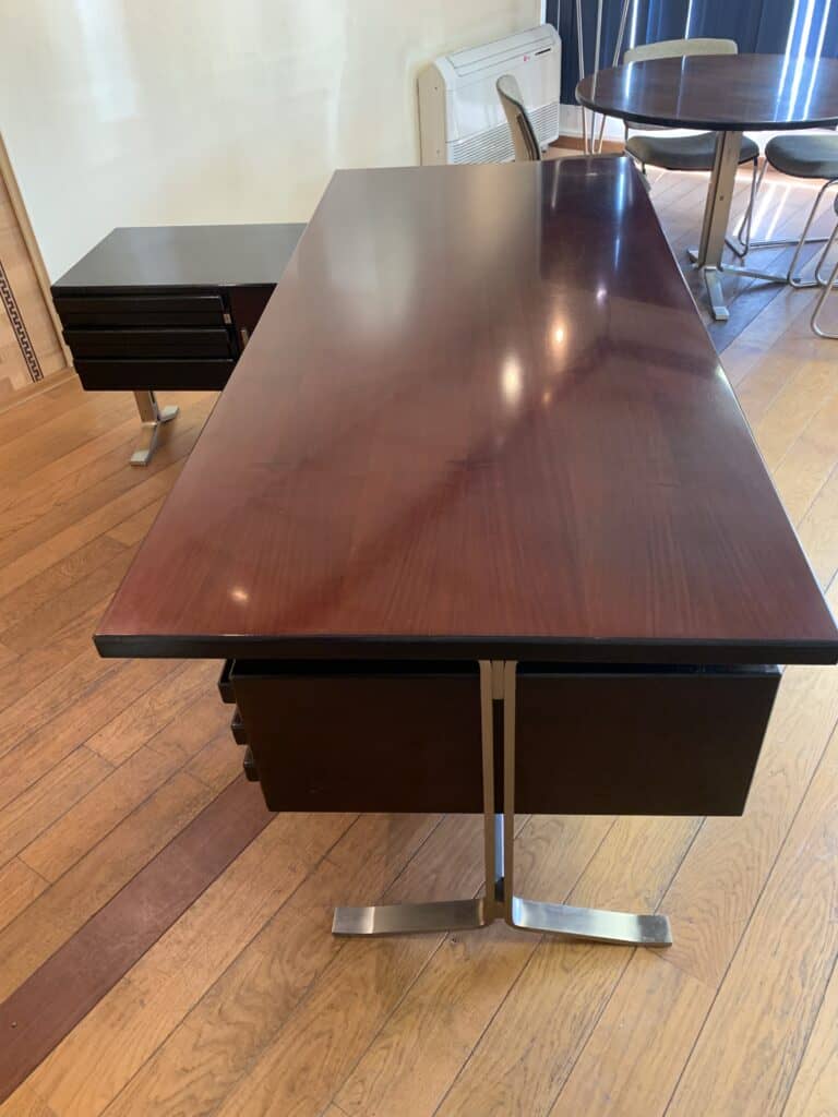 Een gepolijst houten bureau met metalen poten en lades staat op een houten vloer. Op de achtergrond staan nog een bijpassende bijzettafel en een ronde tafel met stoelen.