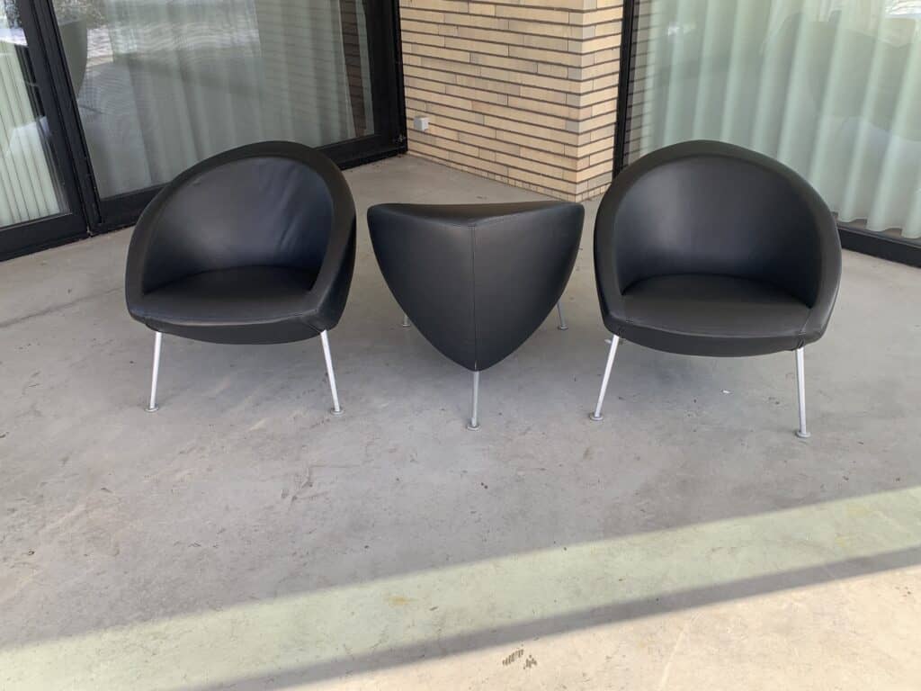 Twee moderne zwarte fauteuils met metalen poten staan op een betonnen terras, tegenover een kleine driehoekige zwarte tafel. Daarachter bevinden zich grote ramen met lichtgordijnen en een bakstenen muur.