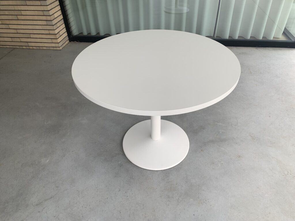 Een ronde, witte tafel in de vorm van een voetstuk staat op een grijze betonnen vloer, voor een glazen wand met verticale jaloezieën en een hoek van baksteen.