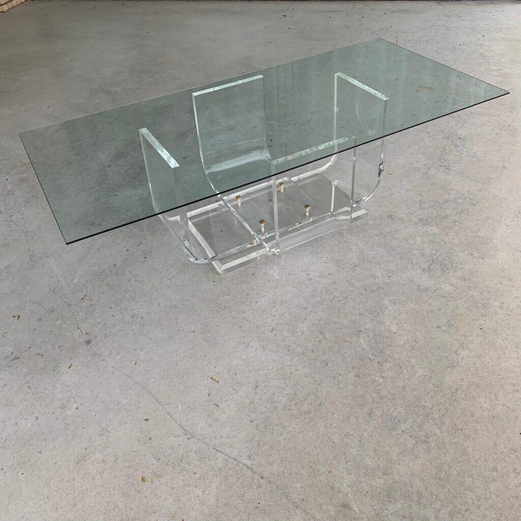 Een moderne, rechthoekige tafel met glazen blad, een onderstel van helder acryl en goudkleurige verbindingsstukken, geplaatst op een eenvoudige betonnen vloer.