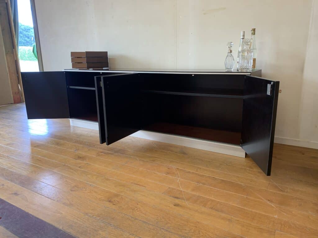Een lang, modern zwart dressoir met beide kastdeuren open, waardoor planken zichtbaar zijn. Er staan flessen en een karaf op. Het meubilair staat op een houten vloer tegen een lichtgekleurde muur.