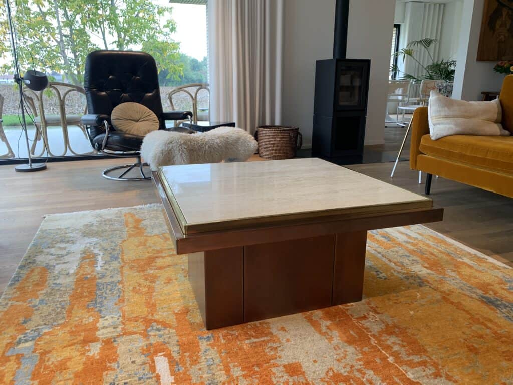 De moderne woonkamer is voorzien van een vierkante salontafel met marmeren blad en een oranje en beige tapijt. Op de achtergrond staan een zwarte schommelstoel, een gouden bank en een houtkachel bij grote ramen.