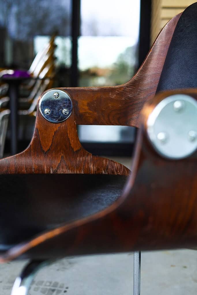 Close-up van een moderne houten stoel met metalen accenten, waarbij de gebogen armleuning en een deel van de zwarte zitting zichtbaar zijn, tegen een onscherpe achtergrond van tafels, stoelen en grote ramen.