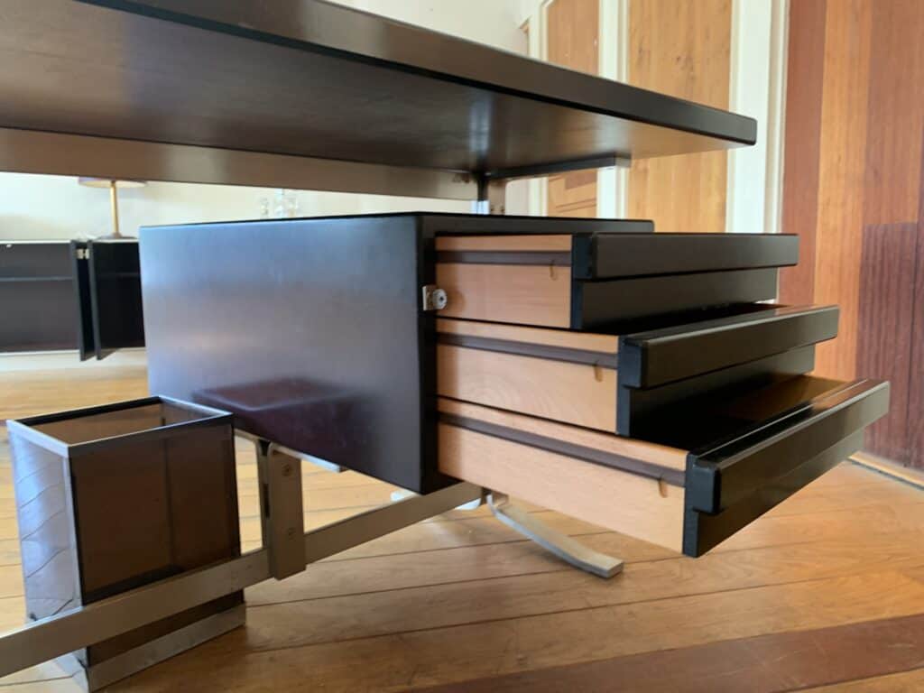 Close-up van een modern bureau met drie halfopen houten lades en daaronder een prullenbak. Dit alles staat op een houten vloer in een goed verlichte kamer met houten deuren en witte muren.