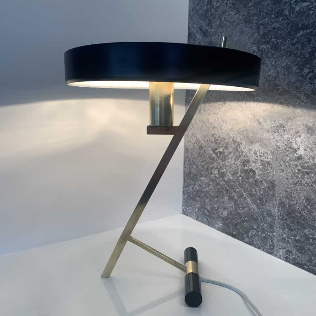 Een moderne bureaulamp met een zwarte, ronde kap, een hoekige standaard van goud en messing en een cilindrische voet, geplaatst op een wit oppervlak met een grijze muur met structuur op de achtergrond.