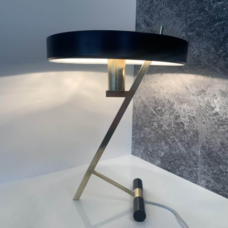 Een moderne bureaulamp met een zwarte, ronde kap, een hoekige standaard van goud en messing en een cilindrische voet, geplaatst op een wit oppervlak met een grijze muur met structuur op de achtergrond.