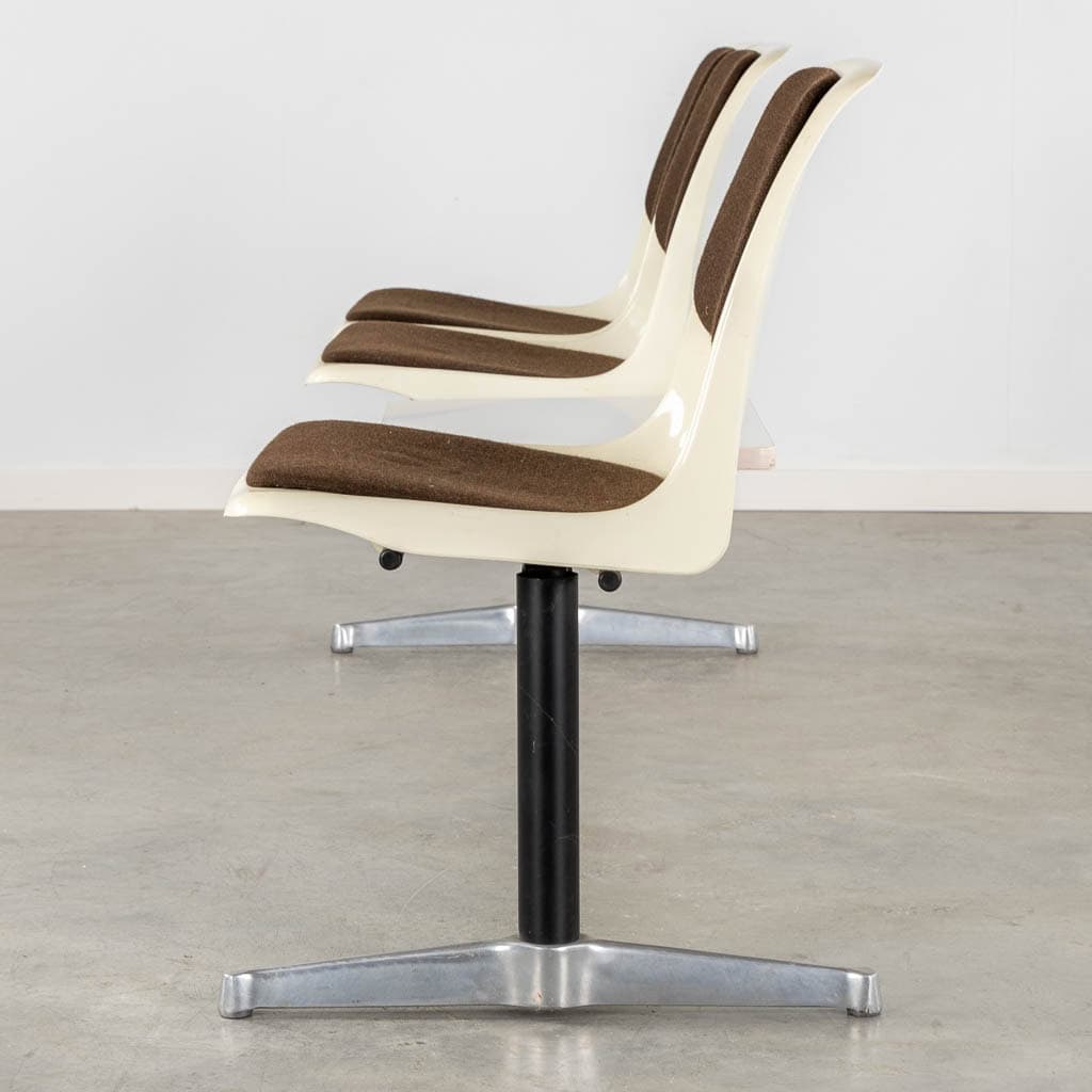 Een rij van drie aaneengeschakelde witte plastic stoelen met bruine zit- en rugleuningkussens, ondersteund door een enkele zwarte metalen voet. Ze staan op een grijze vloer tegen een effen witte muur.