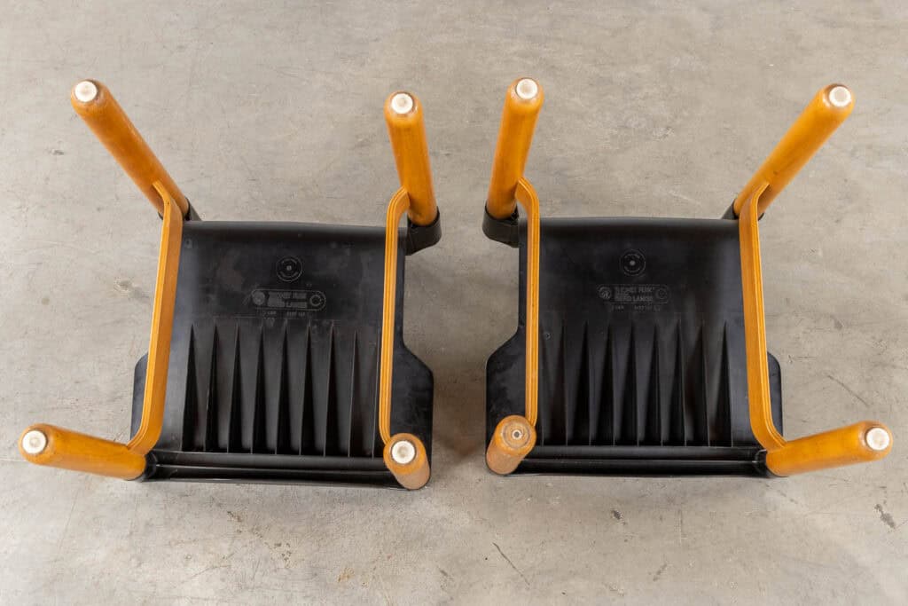 Twee houten stoelen staan ondersteboven op een betonnen vloer, waardoor de zwarte plastic onderkant en de vier poten zichtbaar zijn. De stoelen staan naast elkaar met de poten omhoog gericht.