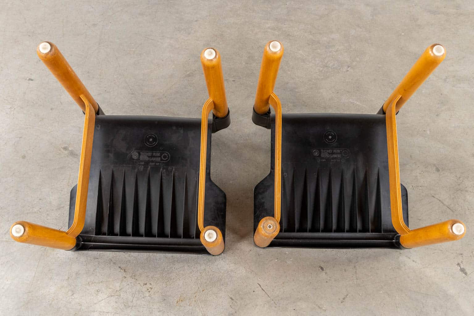 Twee houten stoelen staan ondersteboven op een betonnen vloer, waardoor de zwarte plastic onderkant en de vier poten zichtbaar zijn. De stoelen staan naast elkaar met de poten omhoog gericht.