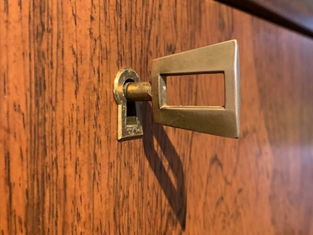 Een close-up van een messing sleutel in een sleutelgat op een houten oppervlak, mogelijk een kast of lade. De sleutel heeft een kenmerkende driehoekige boog met een rechthoekige uitsparing.
