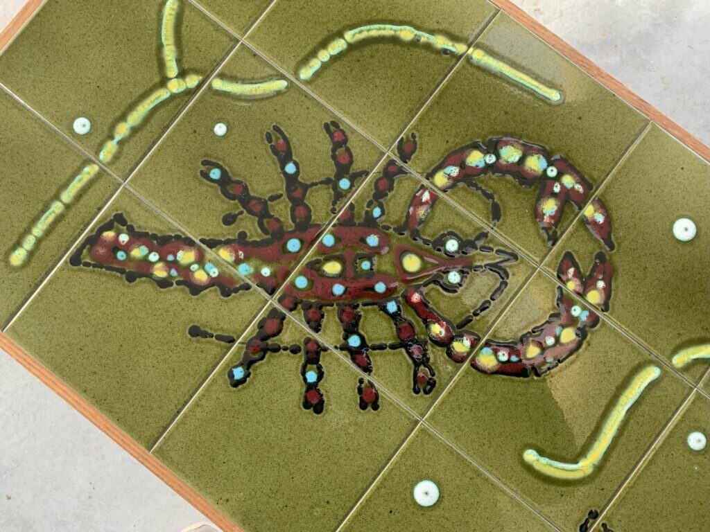 Een tafelblad van keramische tegels met een kleurrijk, abstract kreeftenmotief met groene, rode en blauwe accenten op een olijfkleurige achtergrond, omgeven door golvende gele lijnen en kleine cirkels.