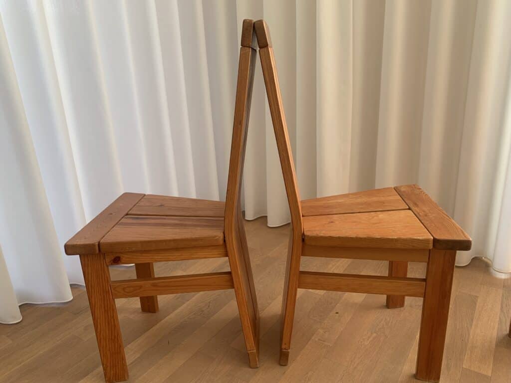 Twee houten stoelen staan rug aan rug, waardoor een symmetrische opstelling ontstaat. De stoelen staan op een lichte houten vloer met witte gordijnen op de achtergrond.