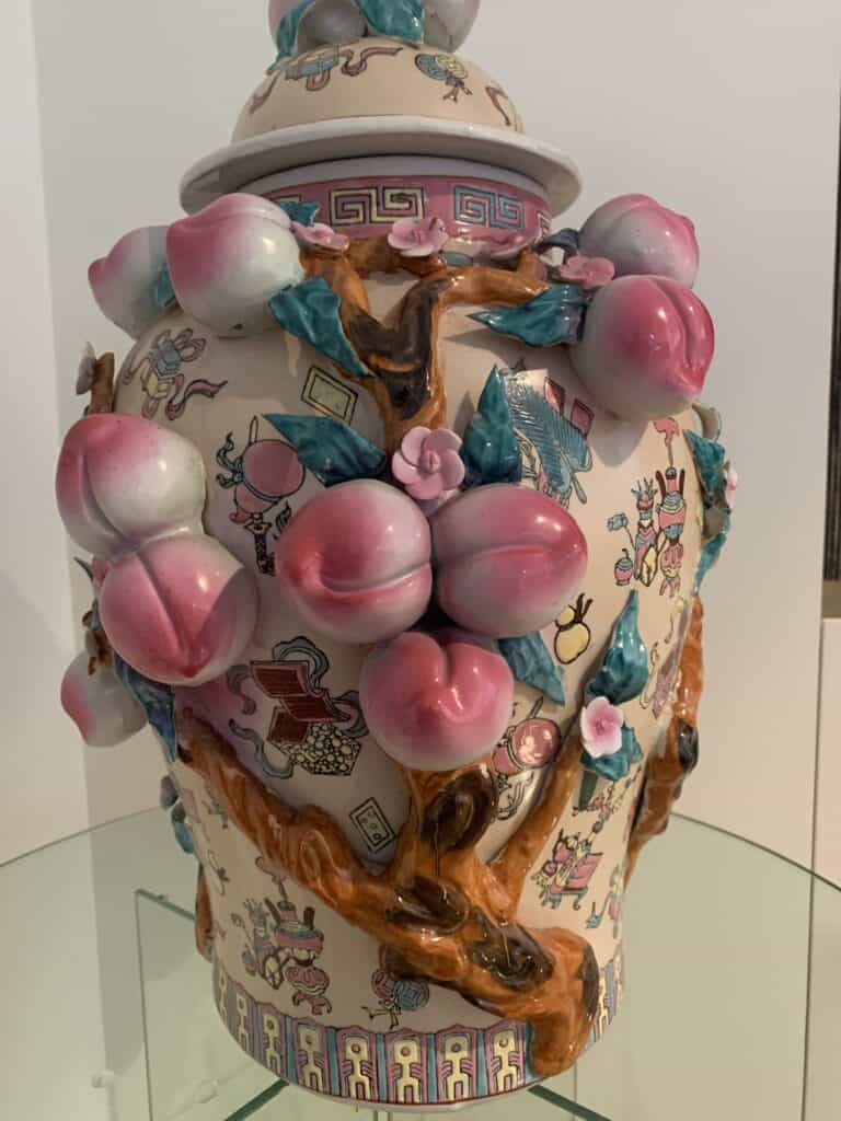 Een decoratieve keramische vaas met deksel, versierd met roze en groene perziksculpturen en kronkelende takken, met ingewikkelde kleurrijke illustraties en patronen op een witte achtergrond. De vaas staat op een glazen oppervlak.