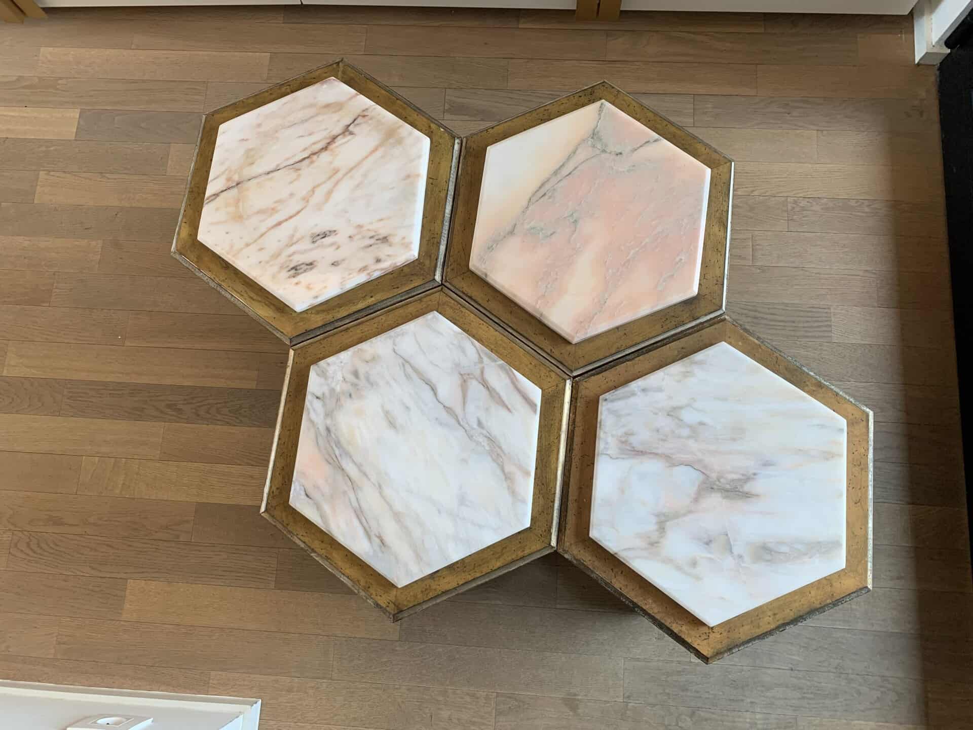 Vier zeshoekige tafels met marmeren blad en messing randen staan naast elkaar op een houten vloer en vormen een honingraatpatroon. De marmeren oppervlakken hebben lichtroze en witte tinten met subtiele adering.