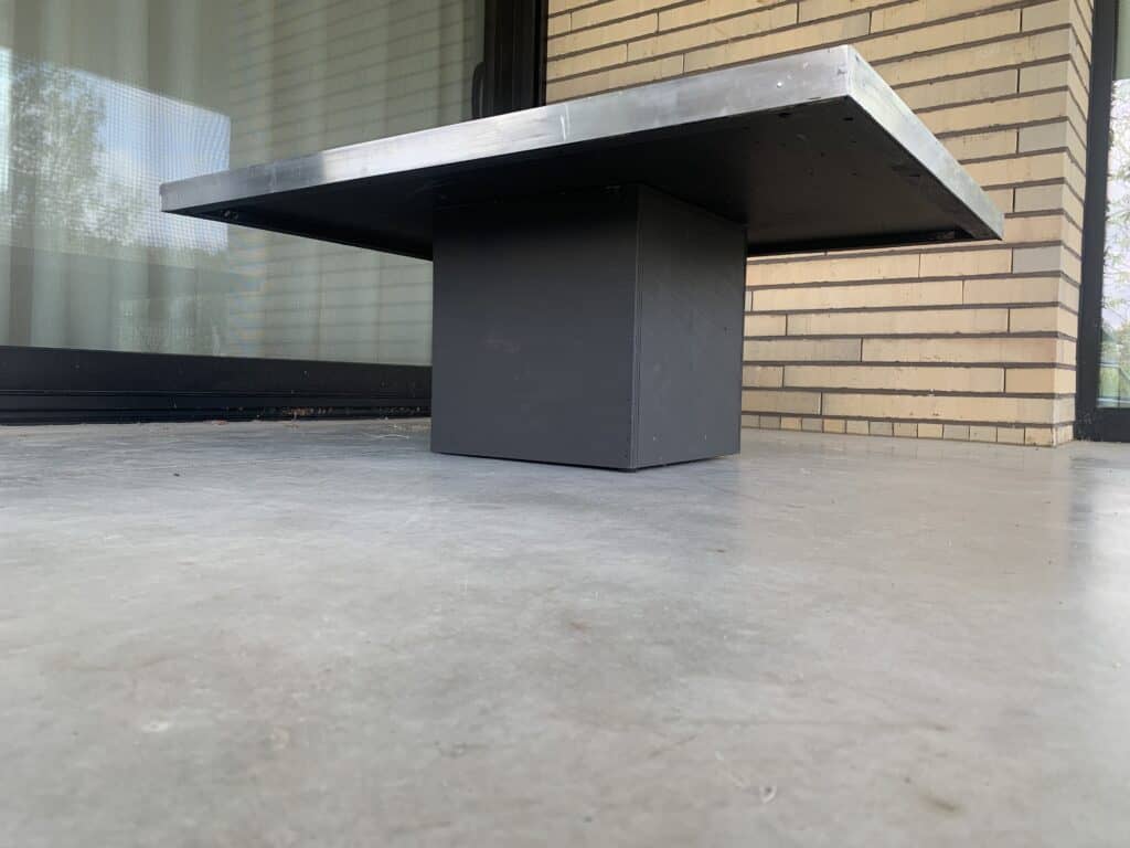 Een lage, moderne tafel met een dik, vierkant metalen blad en een rechthoekig, massief zwart onderstel staat op een betonnen terras bij een bakstenen muur en een glazen schuifdeur.