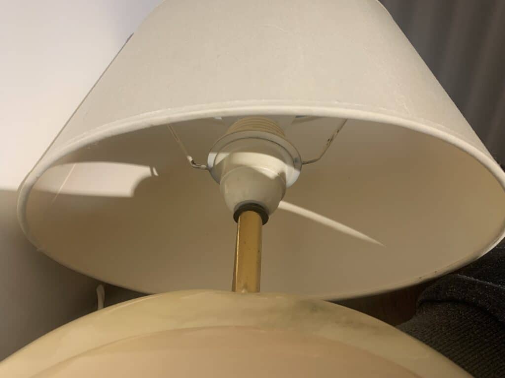 Een close-up van een lamp met een beige lampenkap, waarbij de fitting en de lampenstandaard van onder de kap zichtbaar zijn. Op de voorgrond is een deel van een andere lamp of een ander object zichtbaar.