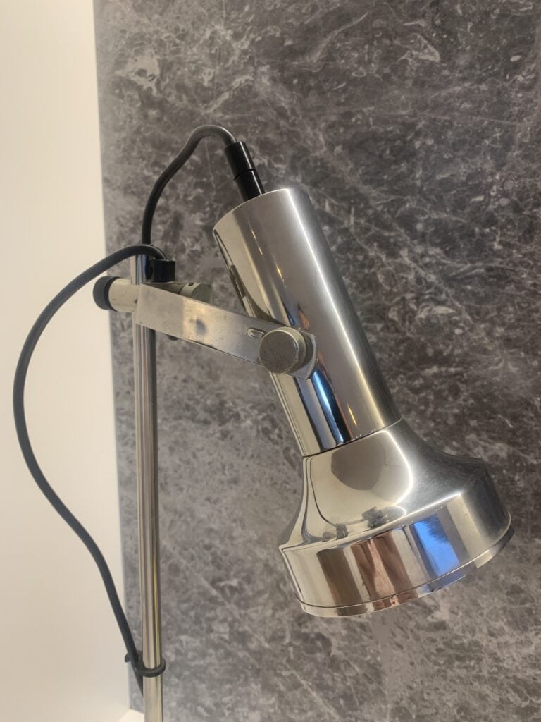 Close-up van een metalen bureaulamp met een cilindrische kap, gemonteerd op een verstelbare arm, tegen een grijze, gemarmerde achtergrond. Het netsnoer is zichtbaar aan de bovenkant van de lamp.