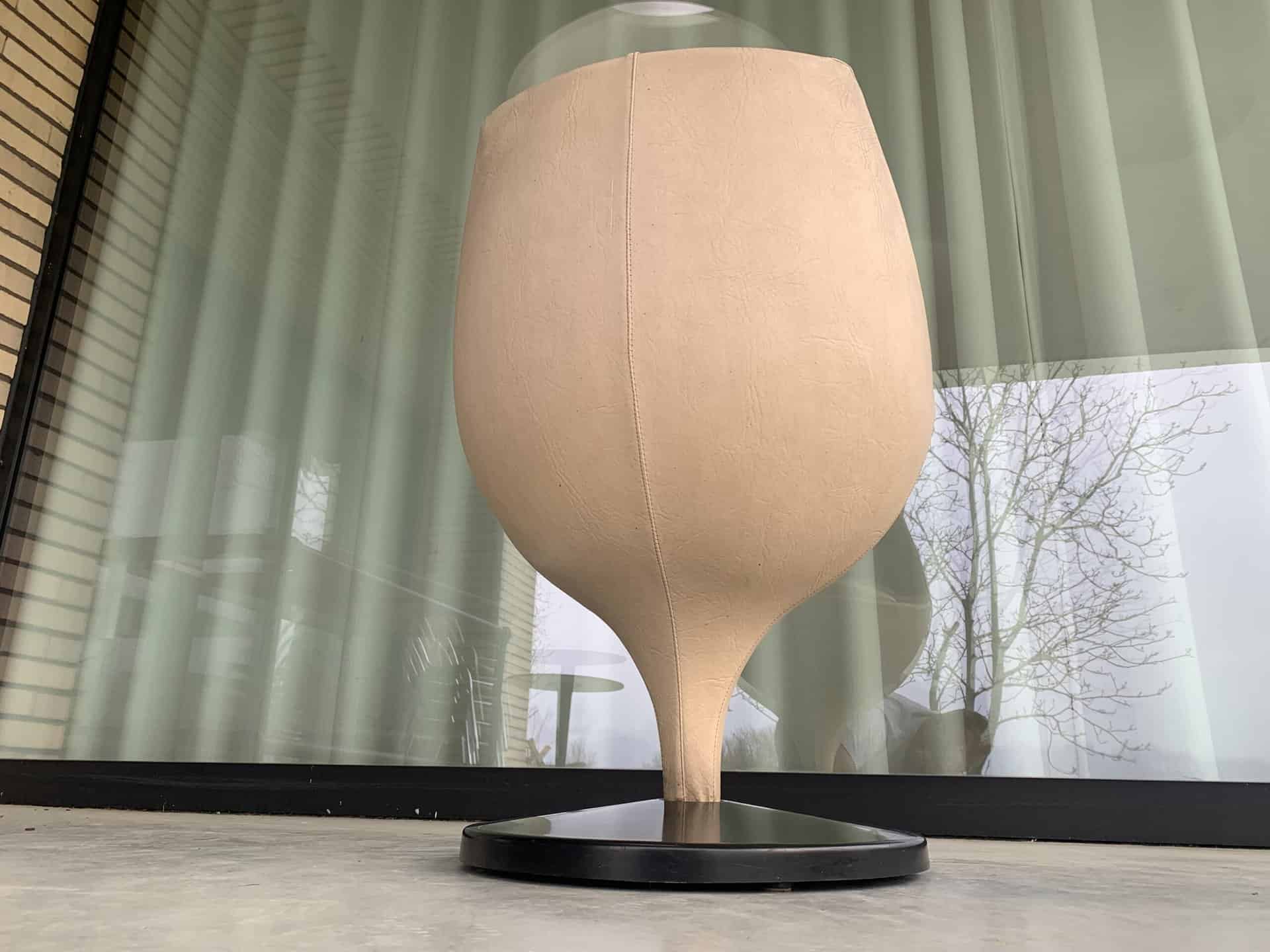 Een grote, beige, kelkvormige sculptuur met een getextureerd oppervlak staat op een zwarte ronde voet voor een glazen raam met verticale jaloezieën. Op de achtergrond zijn weerspiegelingen en boomtakken te zien.
