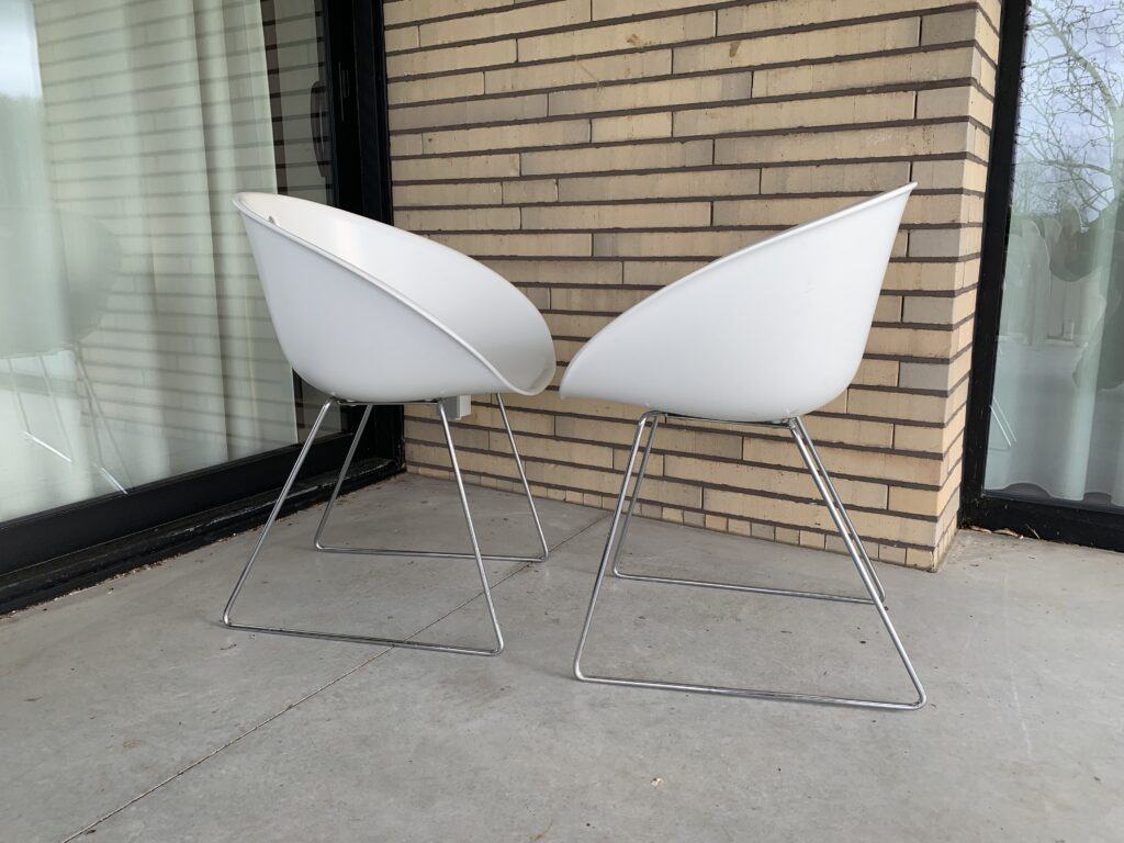 Twee moderne witte stoelen met metalen poten staan tegenover elkaar op een betonnen terras, naast een bakstenen muur en een groot glazen raam.