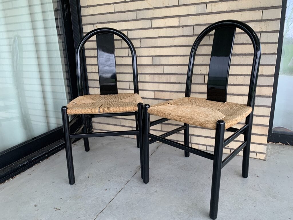 Twee stoelen met zwarte frames en gevlochten rieten zittingen staan naast elkaar op een grijze betonnen vloer, voor een bakstenen muur en een groot raam met vitrage.