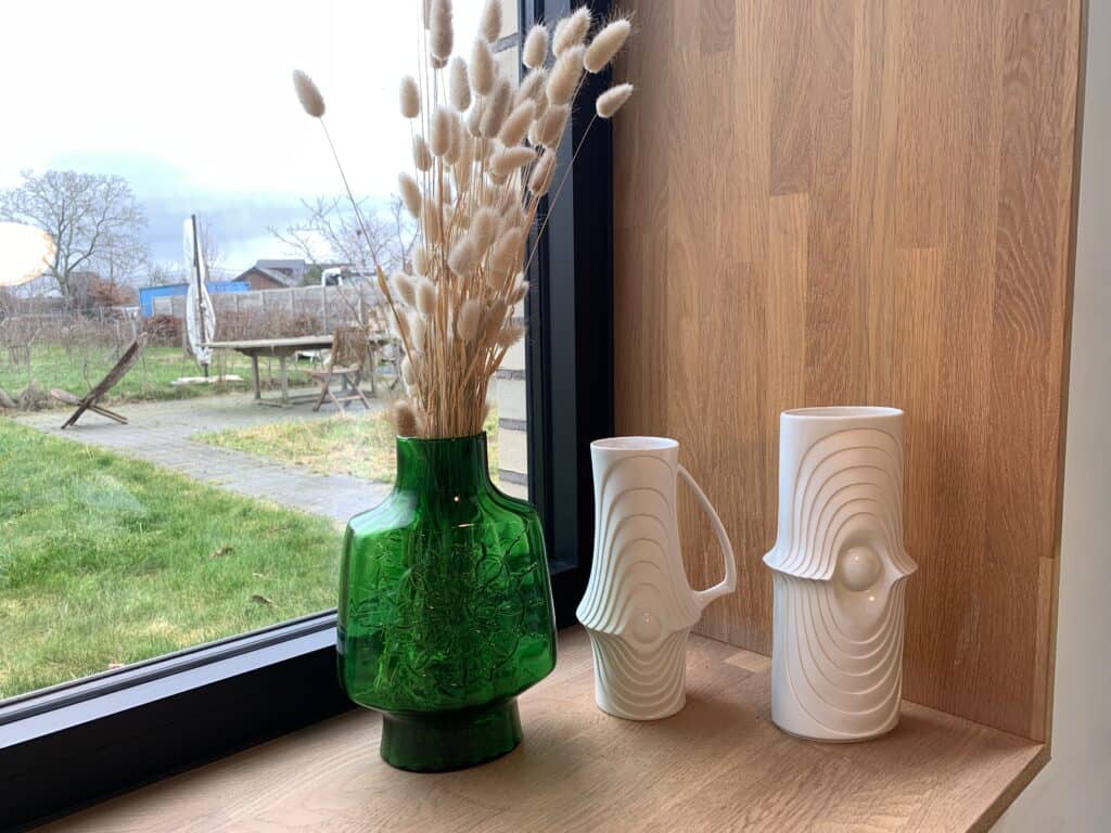 Een groene glazen vaas met gedroogd gras en twee witte decoratieve keramische vazen staan op een houten vensterbank met uitzicht op een tuin en de bewolkte lucht buiten.