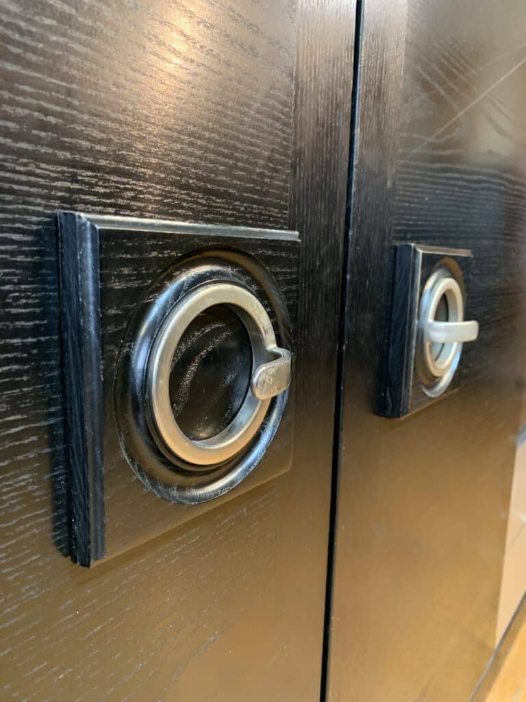 Close-up van twee metalen vierkante handgrepen op een donkere houten kast, met de structuur van de houtnerf en reflecties op de glimmende metalen ringen.