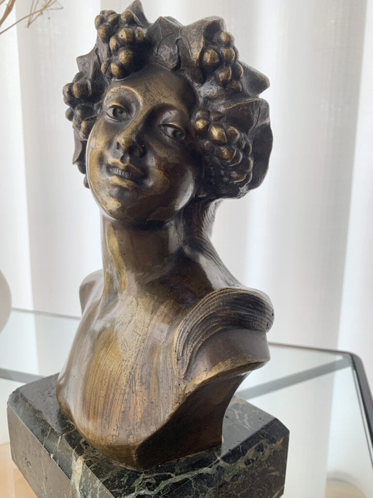 Een bronzen buste van een lachende vrouw met druiventrossen in haar haar, geplaatst op een donkere marmeren voet en tentoongesteld op een glazen oppervlak met witte gordijnen op de achtergrond.