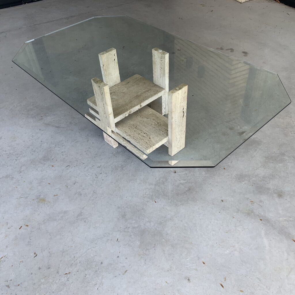 Een glazen salontafel met een onregelmatig achthoekig blad rust op een geometrisch stenen onderstel, geplaatst op een betonnen vloer. Het onderstel bestaat uit gestapelde stenen platen, gerangschikt in een modern, sculpturaal ontwerp.