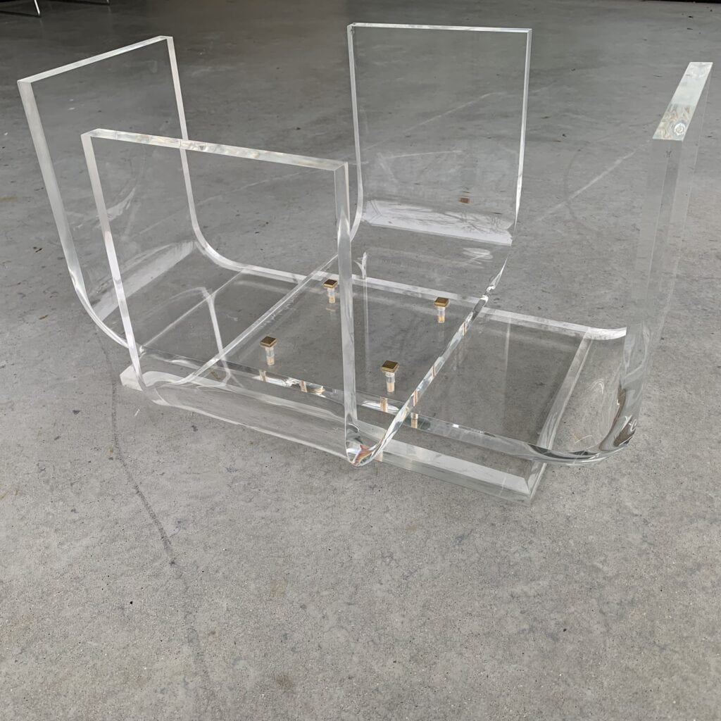 Een doorzichtige acryltafel of tijdschriftenrek ligt ondersteboven op een gladde, grijze betonnen vloer. De transparante, rechthoekige panelen zijn met goudkleurige metalen verbindingsstukken aan elkaar verbonden.