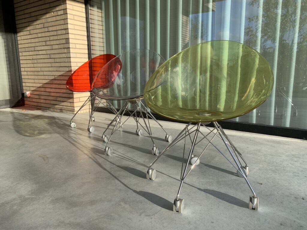 Drie doorschijnende, ronde plastic stoelen in de kleuren rood, transparant en groen staan op een terras met metalen poten en witte wieltjes. Ze staan bij een glazen raam met verticale jaloezieën en een bakstenen muur.