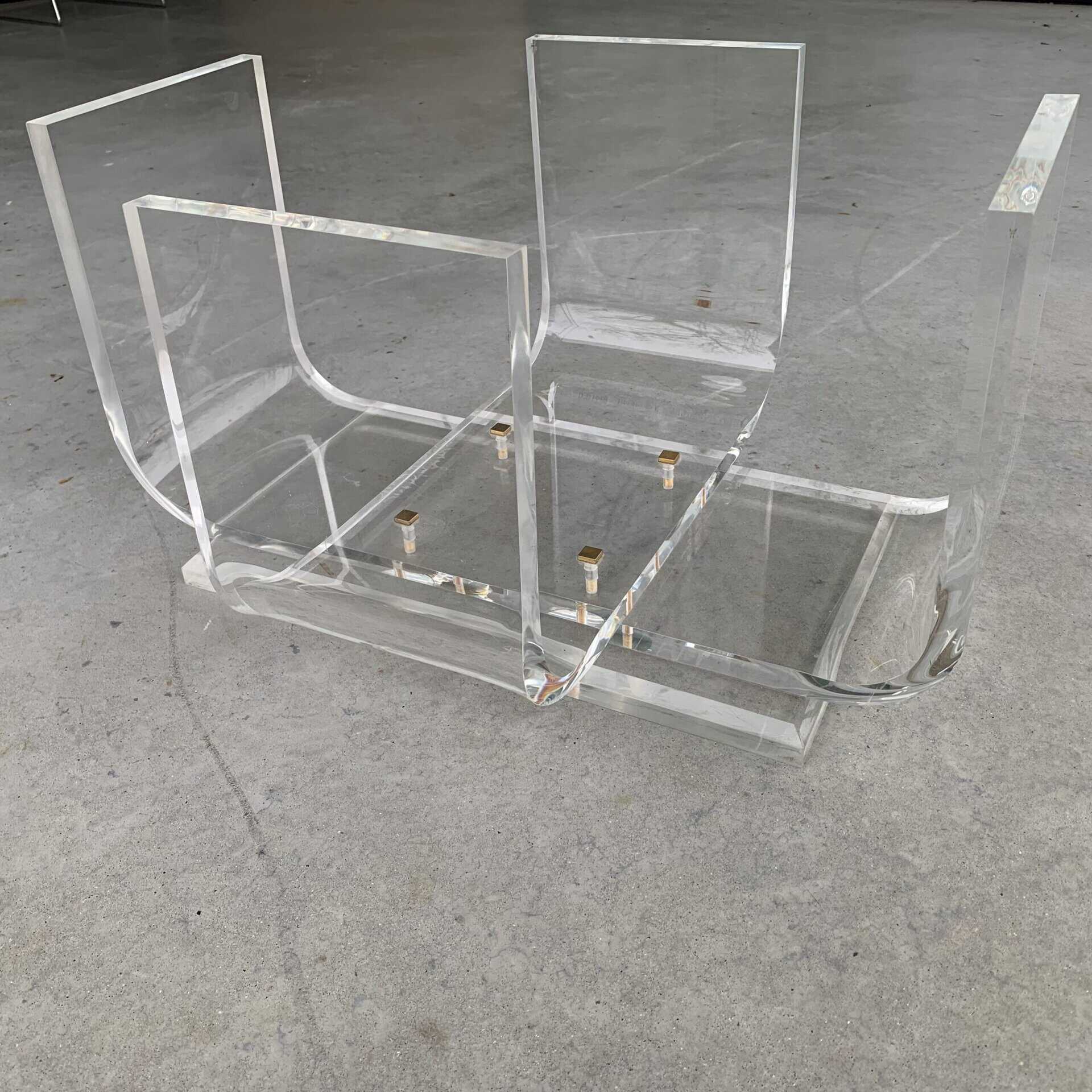 Een doorzichtige acryltafel of tijdschriftenrek ligt ondersteboven op een gladde, grijze betonnen vloer. De transparante, rechthoekige panelen zijn met goudkleurige metalen verbindingsstukken aan elkaar verbonden.