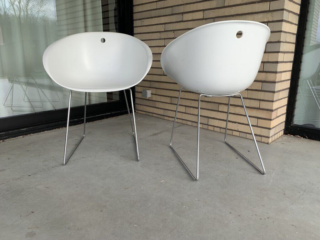 Twee moderne witte stoelen met dunne metalen poten staan op een betonnen terras bij een bakstenen muur en een grote glazen deur. De stoelen hebben afgeronde rugleuningen en een klein rond gat bovenaan.