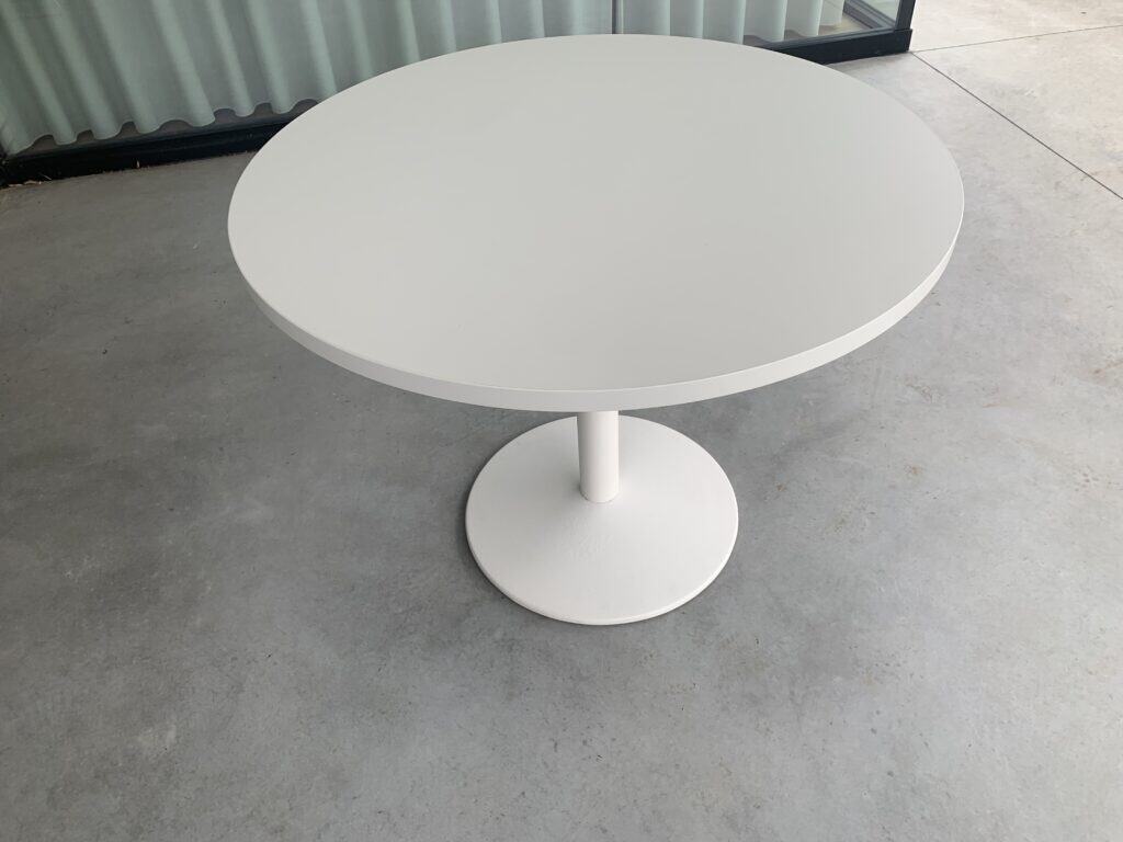 Een ronde, witte tafel met een enkele centrale voet staat op een gladde, lichtgrijze betonnen vloer, vlak bij een glazen raam met verticale jaloezieën.