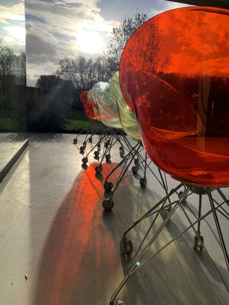 Een rij doorschijnende plastic stoelen in oranje, groen en transparant staat binnen naast een groot raam, waar het zonlicht kleurrijke schaduwen op de vloer werpt. Buiten zijn de bomen en de lucht zichtbaar.