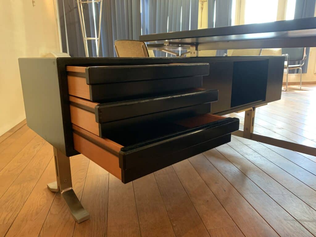 Een close-up van een modern bureau met drie lichtjes openstaande lades, met een afwerking van hout en zwart, staand op metalen poten op een houten vloer in een zonnige kamer met blauwe gordijnen en stoelen op de achtergrond.