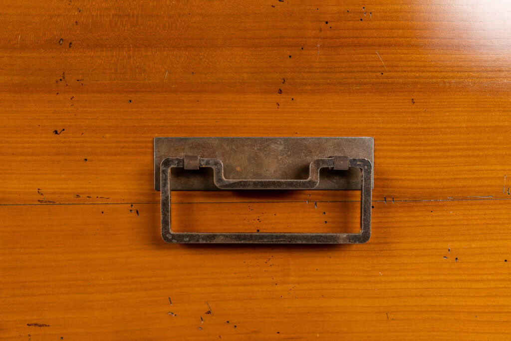 Een rechthoekige metalen ladegreep met een verweerde afwerking is gemonteerd op een houten oppervlak met een warme bruine tint en zichtbare nerfpatronen.