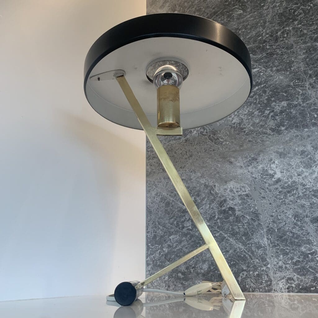 Moderne tafellamp met een ronde, zwarte kap, een hoekige messing standaard en een zichtbare gloeilamp, geplaatst op een wit oppervlak tegen een muur met een gespleten wit-grijze marmerstructuur.